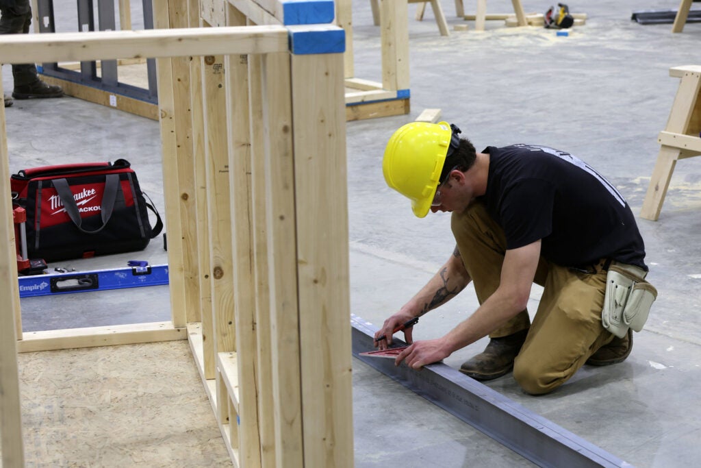 A student in a hard hat measures a metal beam with a triangle, kneeling near a wooden building frame in a construction class at Alfred State College