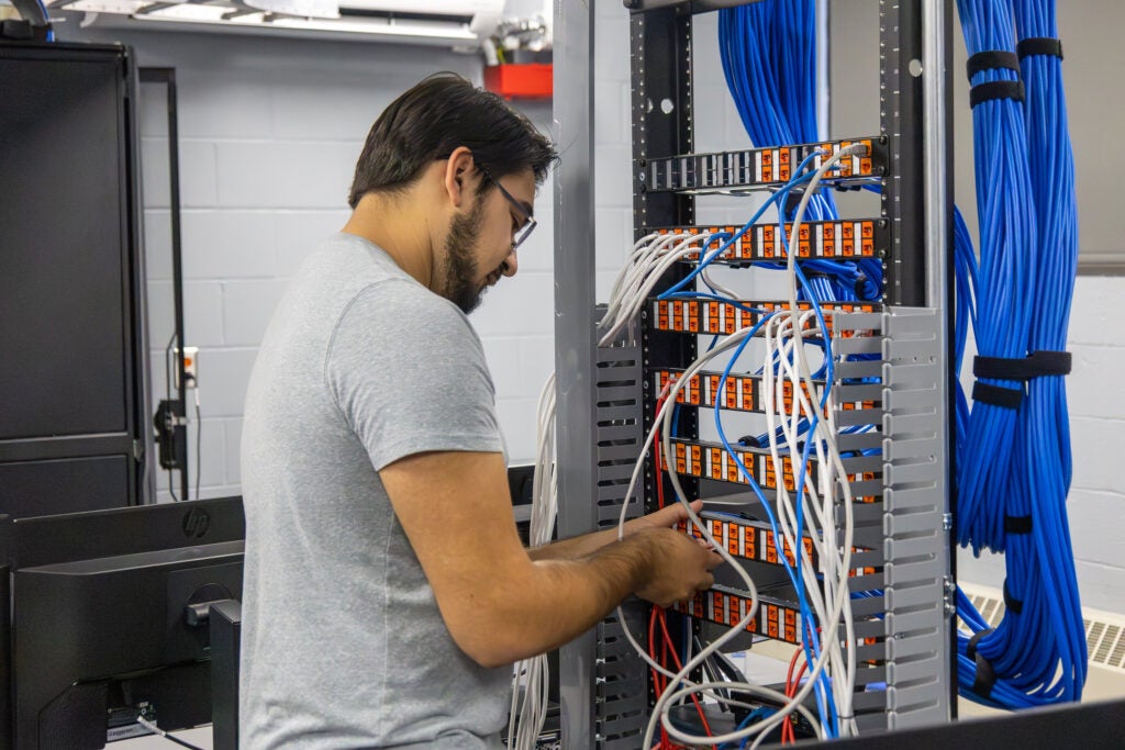 A man with glasses and a beard connects and organizes cables on a patch panel inside a server room.