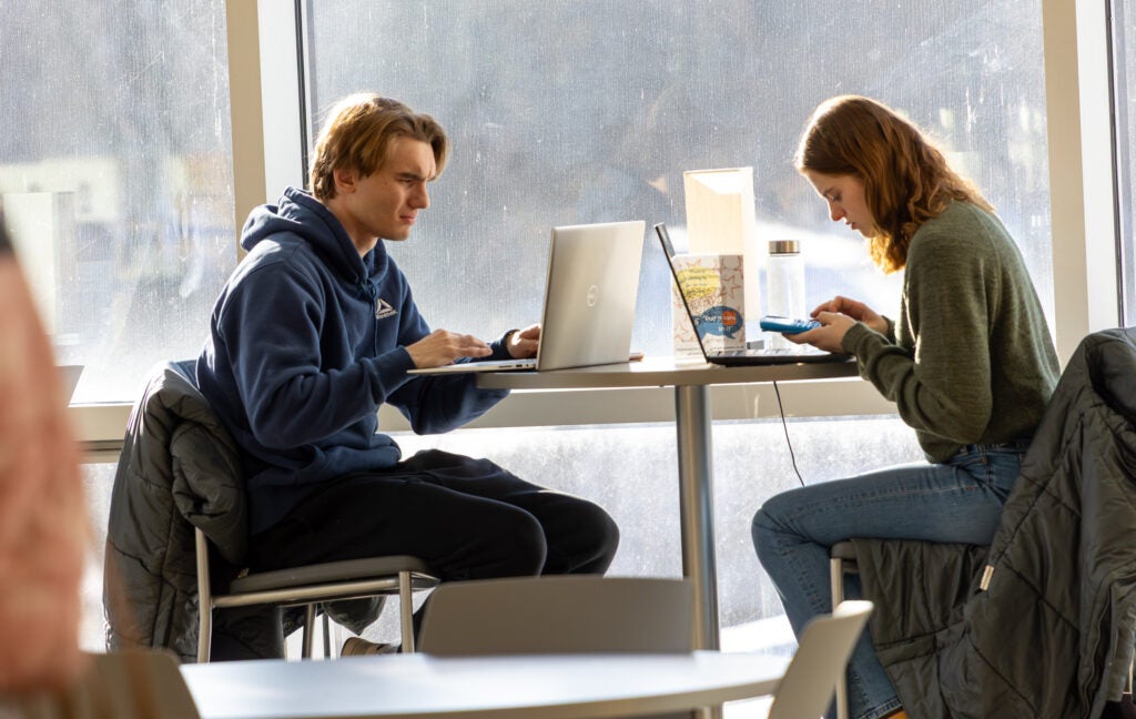 Two students work on laptops near a large window in a common area early in the spring semester at Alfred State College