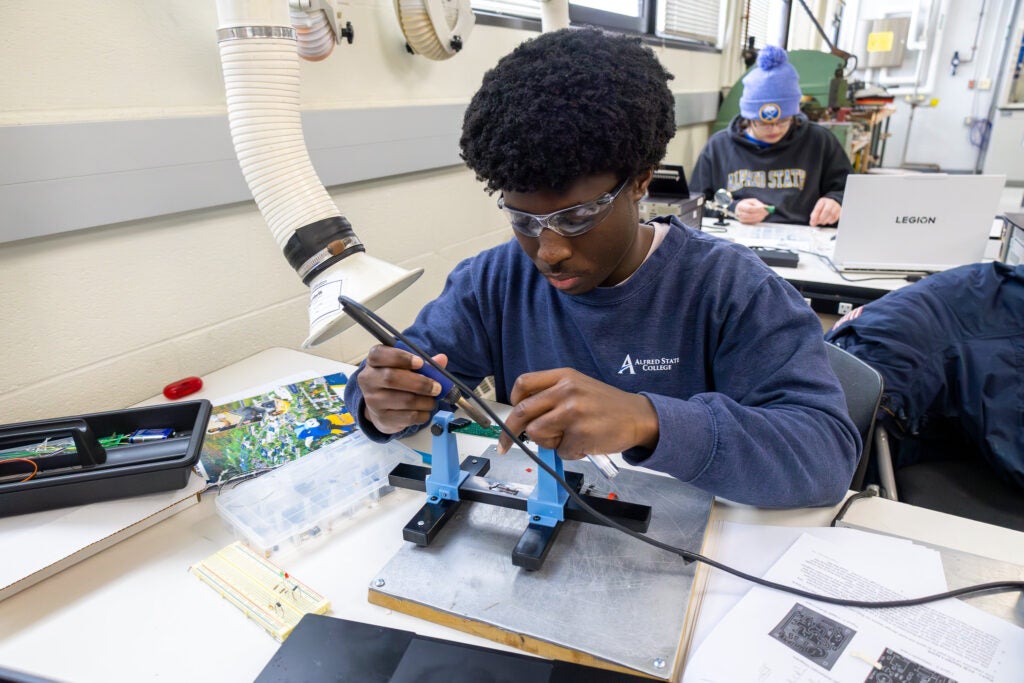 A student wearing safety glasses sits at a desk learning to use electronic fabrication equipment at Alfred State College