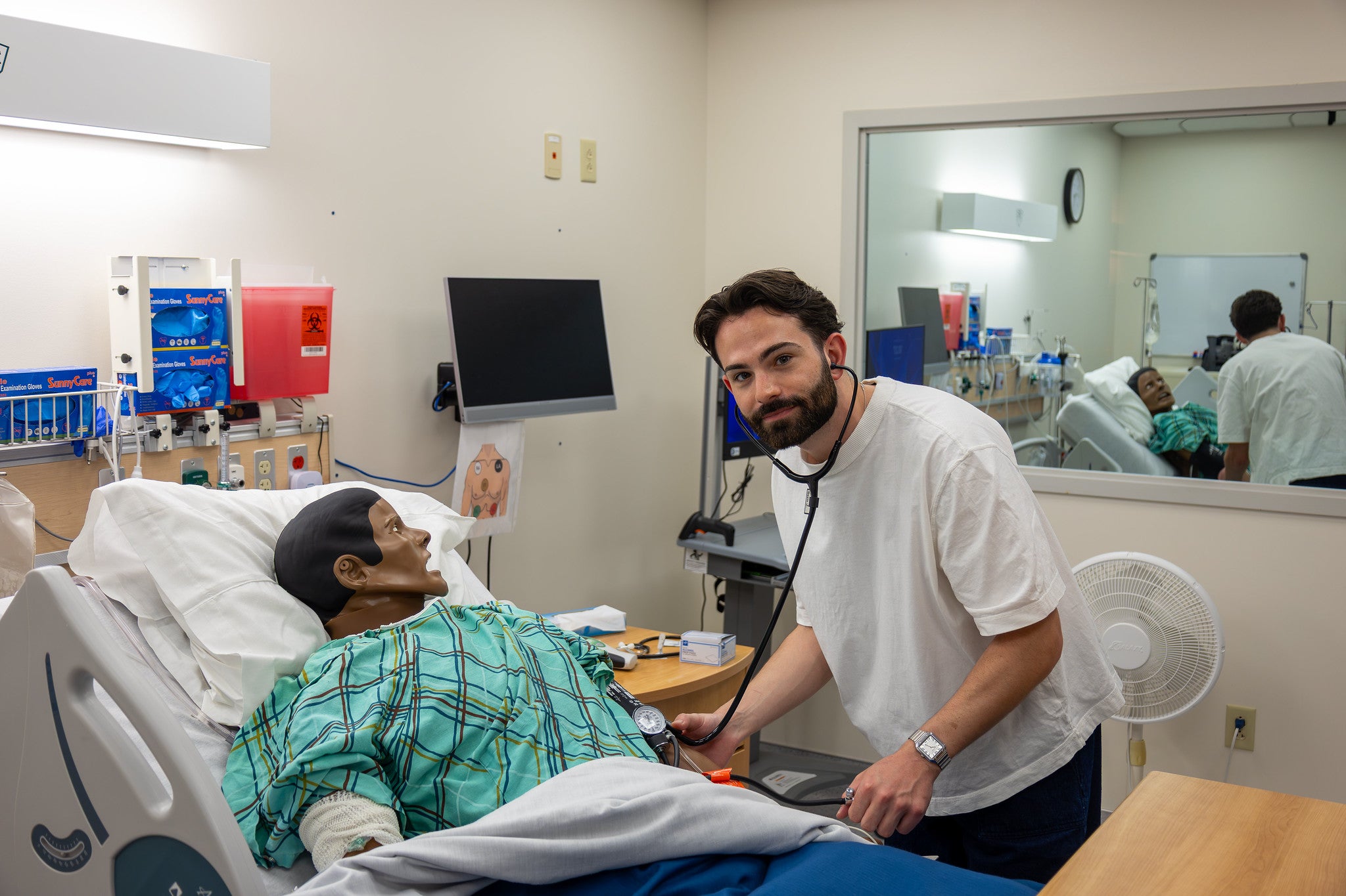 A bearded male medical student wearing a stethoscope smiles at the camera while practicing taking blood pressure on a simulation mannequin lying in a hospital bed inside a training lab.