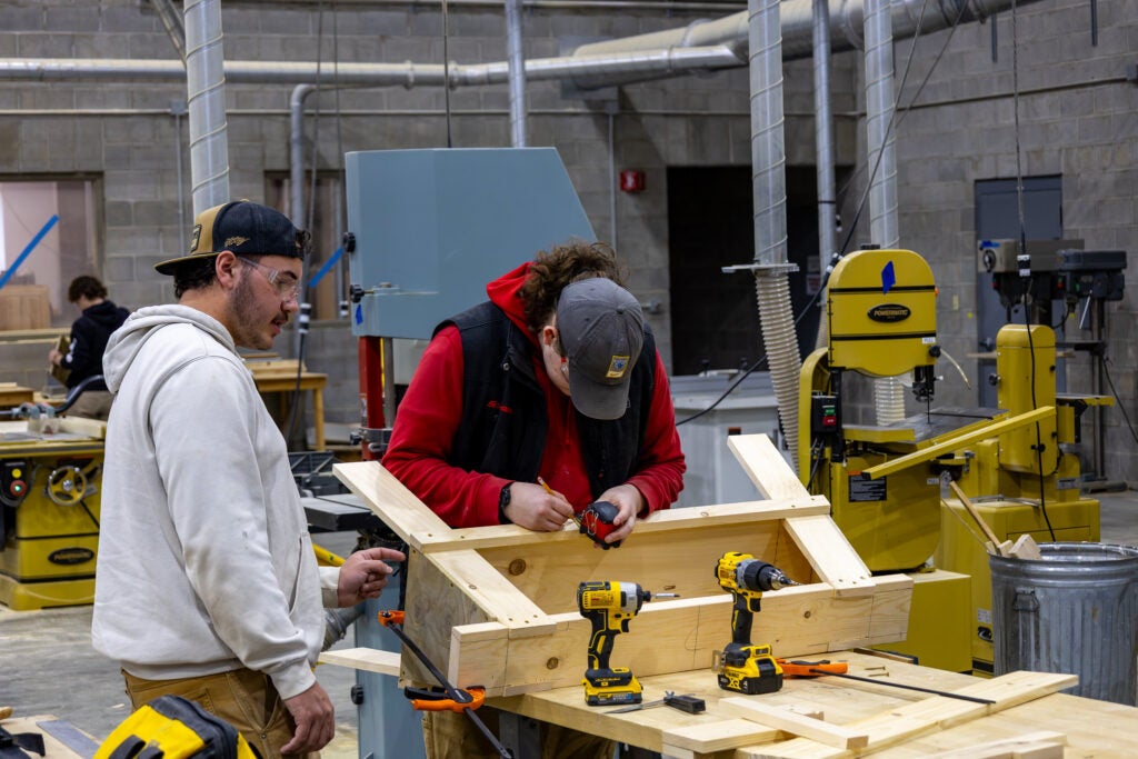 Two students build a wooden platform structure, in a construction space with drills set to the side at Alfred State College