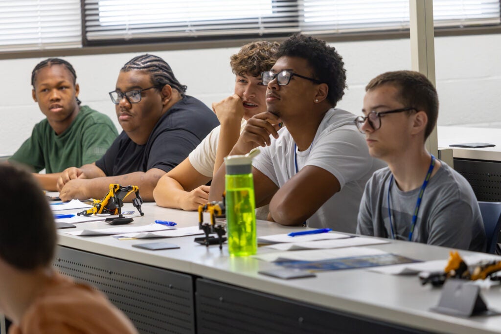 Students sitting along a long desk in a class at Alfred State College listen to a lecture about robotics