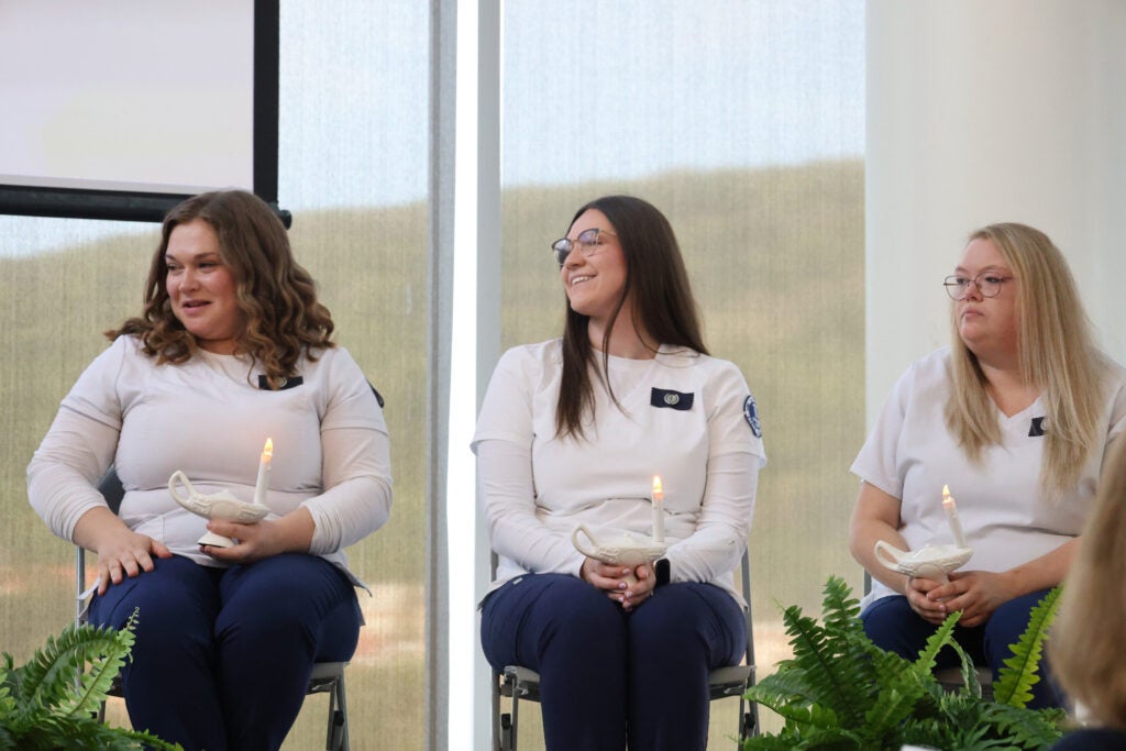 LPN graduates in white and blue sit on stage at Alfred State College holding candles