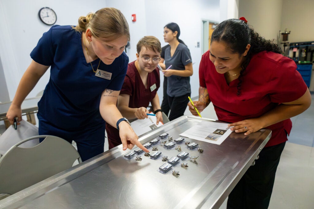 Students in nursing scrubs examine name tags displayed on a metal table in a simulation space at Alfred State College